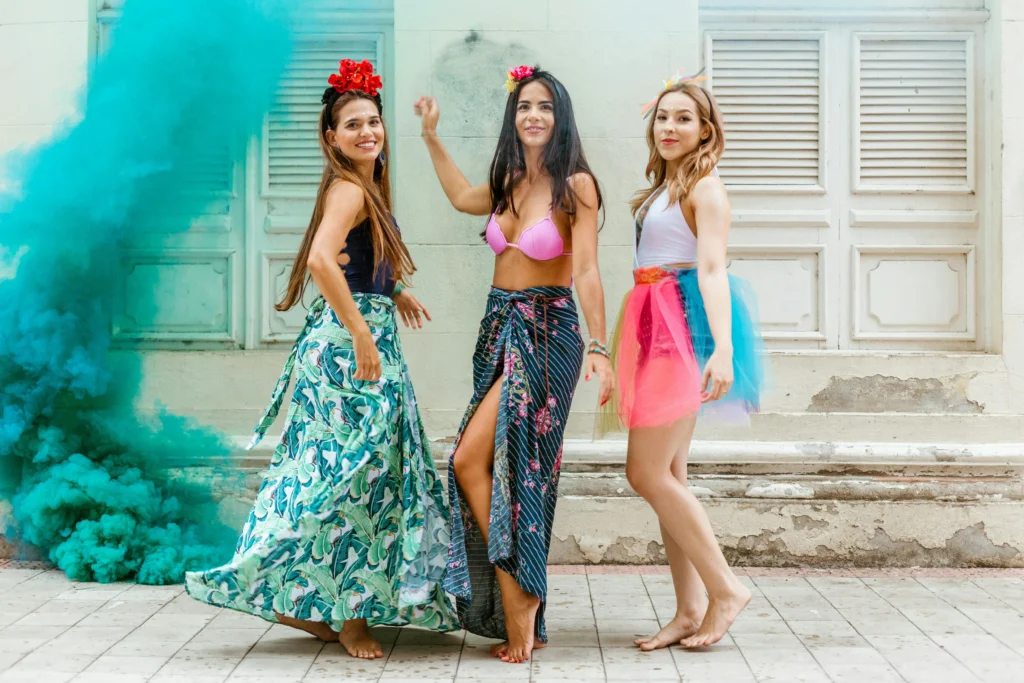 Três mulheres vestidas para o Carnaval em frente a uma casa colonial, com fumaça colorida — representando a alegria e a cultura das festas nordestinas em 2026